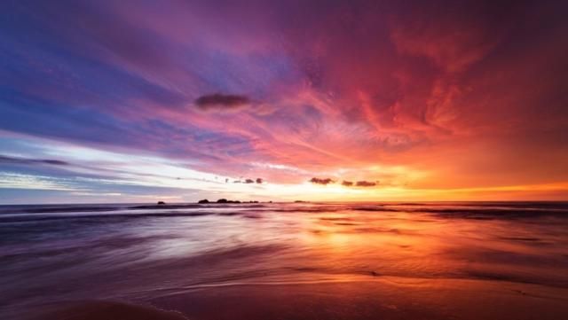 A sunset over the ocean with orange and purple clouds reflected on the wet sand.